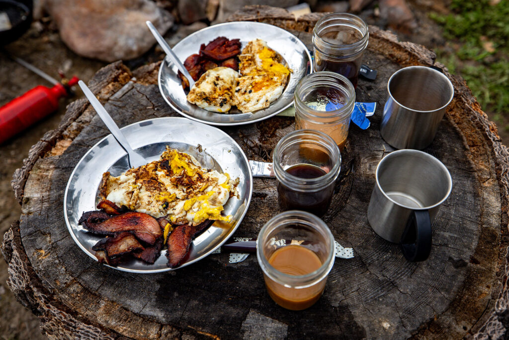 Two plates of eggs and bacon with coffee in glass jars and stainless steel camping mugs arranged on a tree stump outdoors.