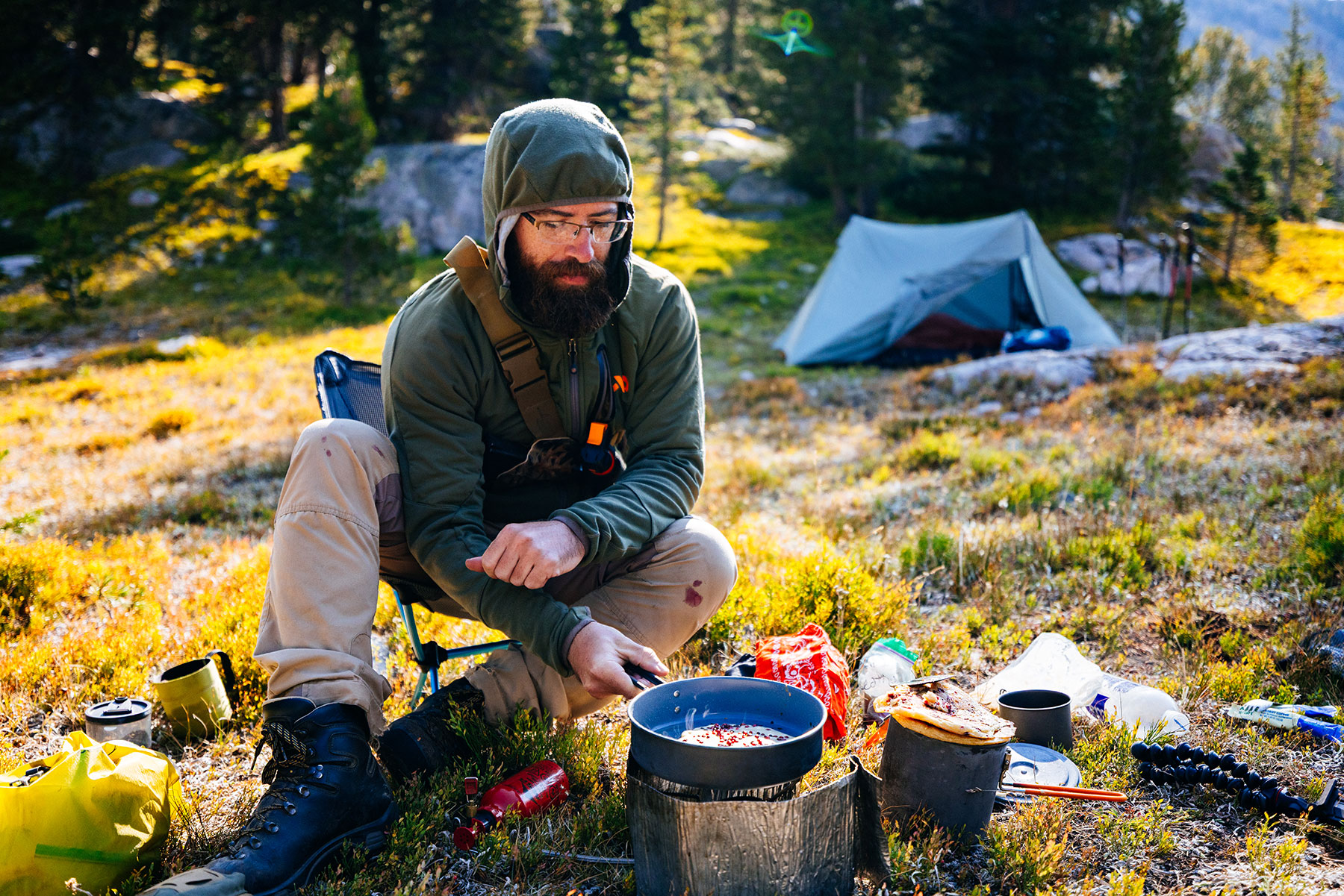 A backpacker cooking breakfast at a backcountry campsite with a lightweight backpacking tent in the background.