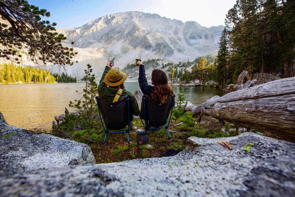 Two backpackers sitting in camp chairs beside an alpine lake, raising their drinks toward the surrounding mountains.