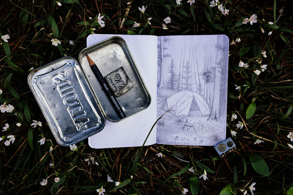 Small sketchbook with a pencil and tin case on the forest floor with a hand-drawn campsite sketch surrounded by small white flowers