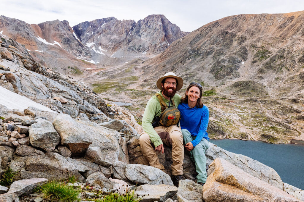 Two hikers sitting on rocky alpine terrain above a mountain lake in Montana, wearing long-sleeve sun shirts, hiking pants, and a sun hat with rugged peaks in the background.