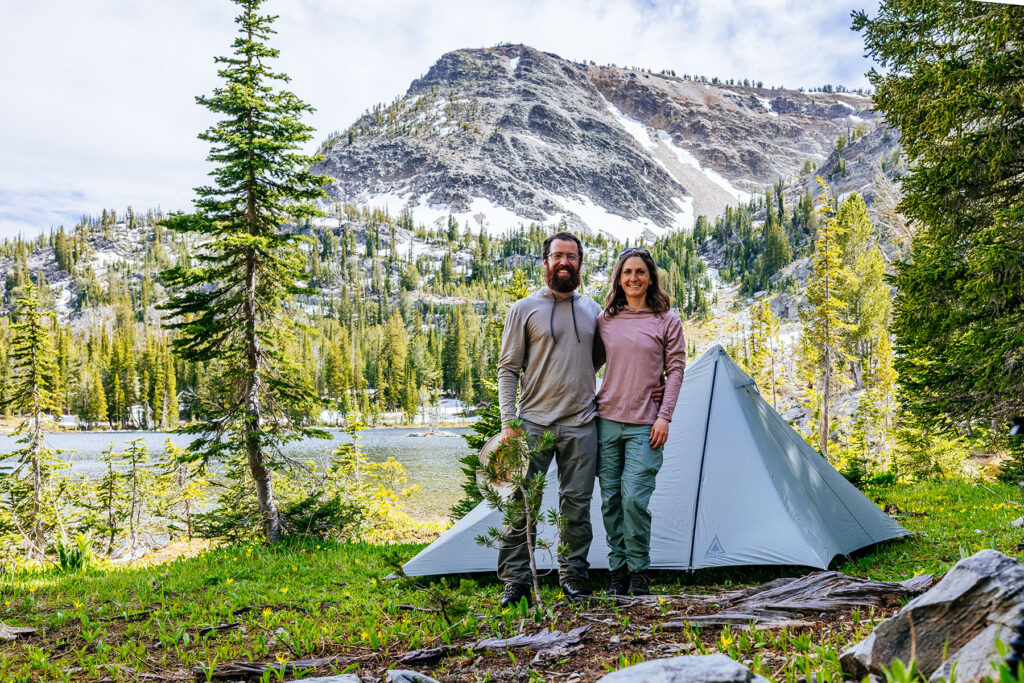 Backyard Montana founders standing beside their Durston Gear X-Mid 2 backpacking tent at an alpine campsite in Montana with a mountain and lake in the background.
