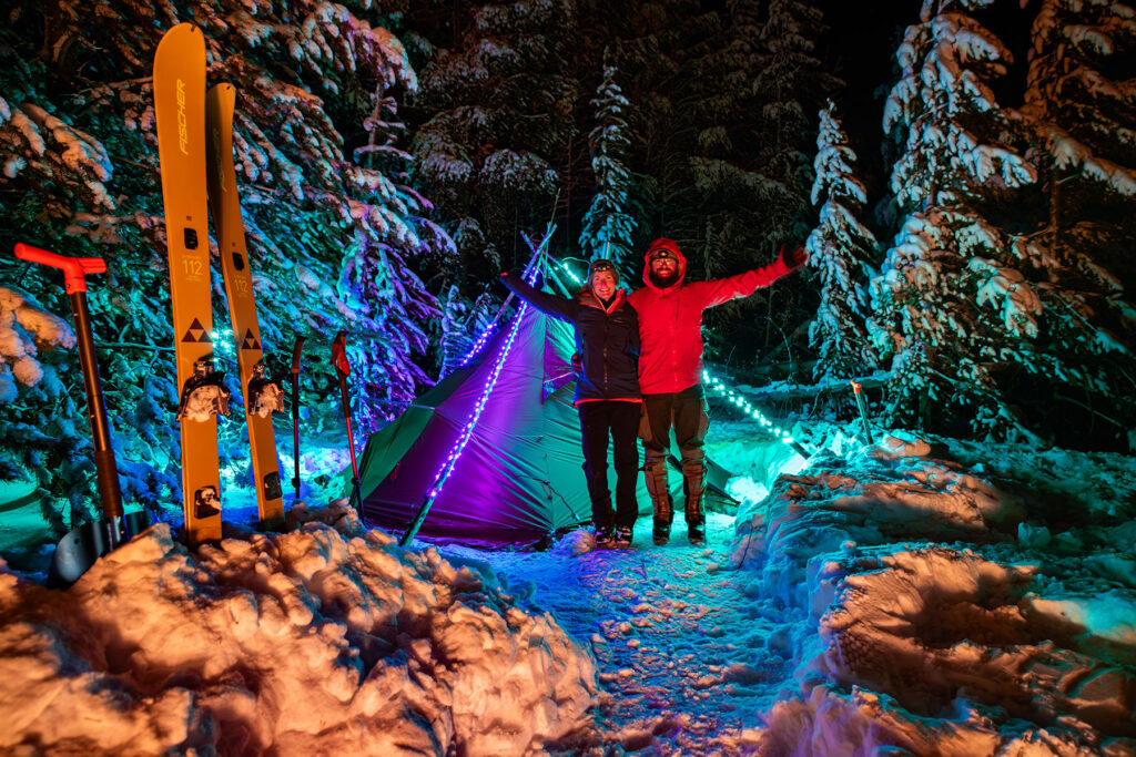 Two winter campers standing beside a tent decorated with colorful string lights in a snowy forest, with skis nearby.