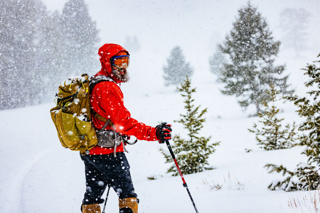 Skier wearing a red shell jacket, goggles, and backpack among evergreen trees during heavy snowfall.