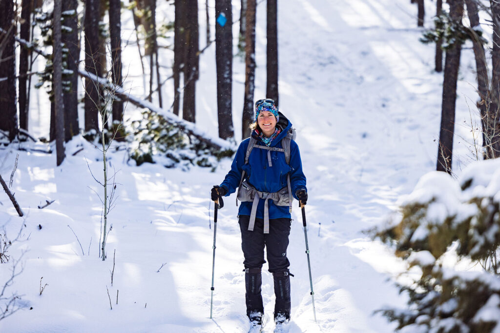Smiling cross-country skier dressed in a blue jacket, winter pants, and backpack on a snow-covered forest trail.