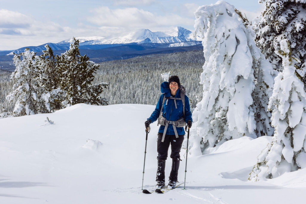 Cross-country skier wearing a blue jacket and backpack on a snowy trail with mountain peaks in the background.