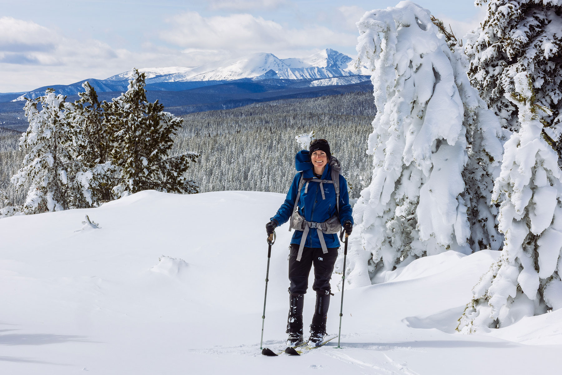 Cross-country skier wearing a blue jacket and backpack on a snowy trail with mountain peaks in the background.
